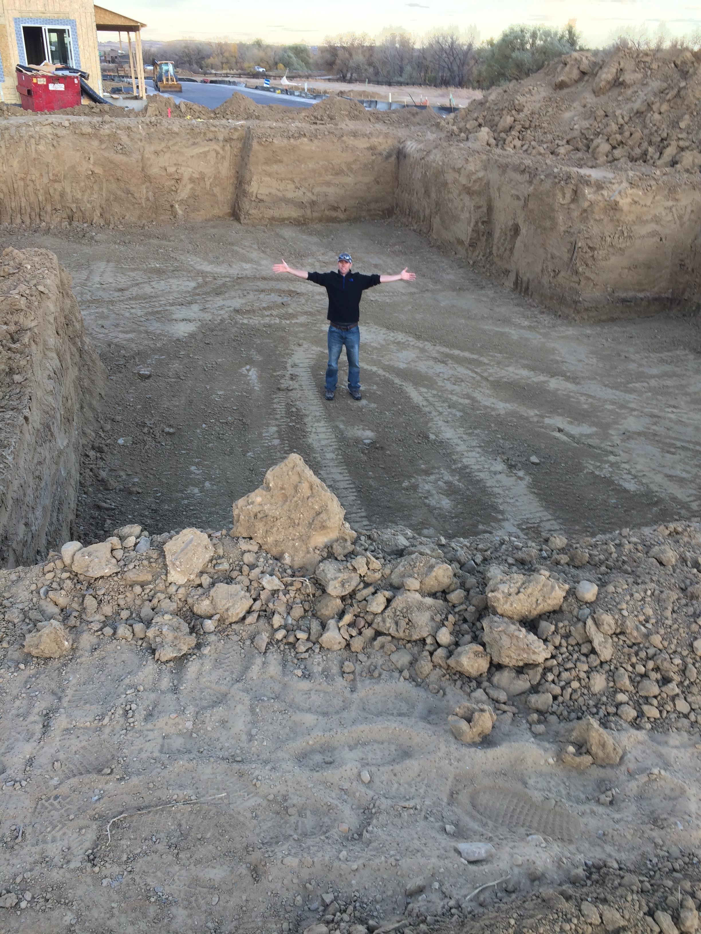 A man standing in the middle of a large excavated lot at a construction site, with arms outstretched, surrounded by piles of dirt and construction equipment in the background.