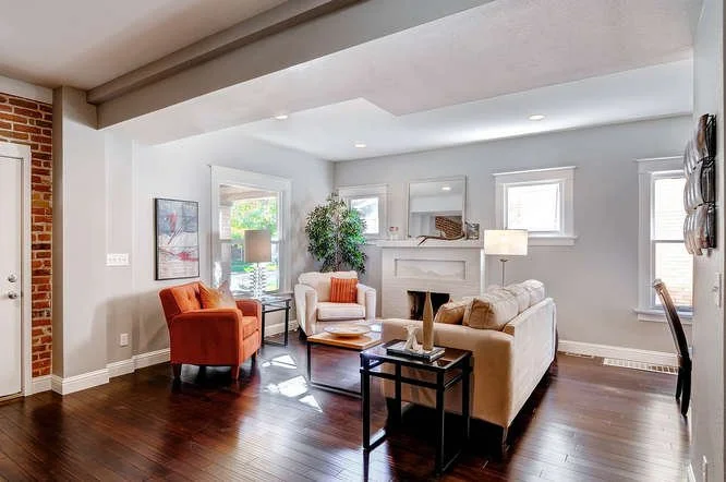 Living room with white walls, hardwood floors, and natural light from windows. There is a white fireplace, a cream sofa with brown pillows, an orange armchair, an off-white armchair, and side tables with lamps. Decor includes wall art, a potted plant, and a mirror.