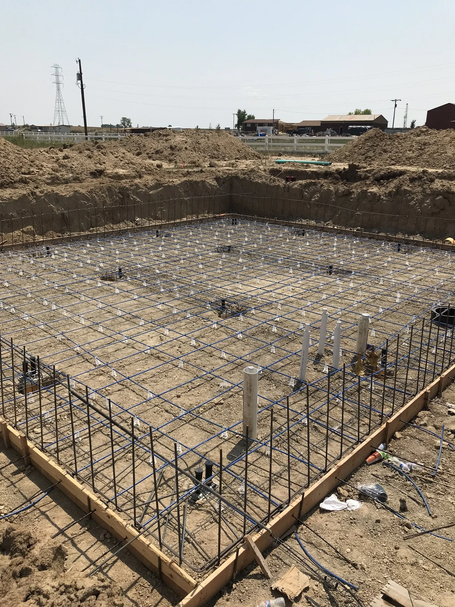 Construction site with a foundation and rebar framework, pipes, and dirt mounds, with buildings and power lines in the background.