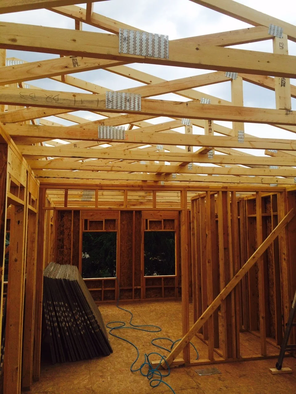 Inside view of a house under construction, showing wooden framing, roof trusses, and an unfinished interior with windows, construction materials, and tools.