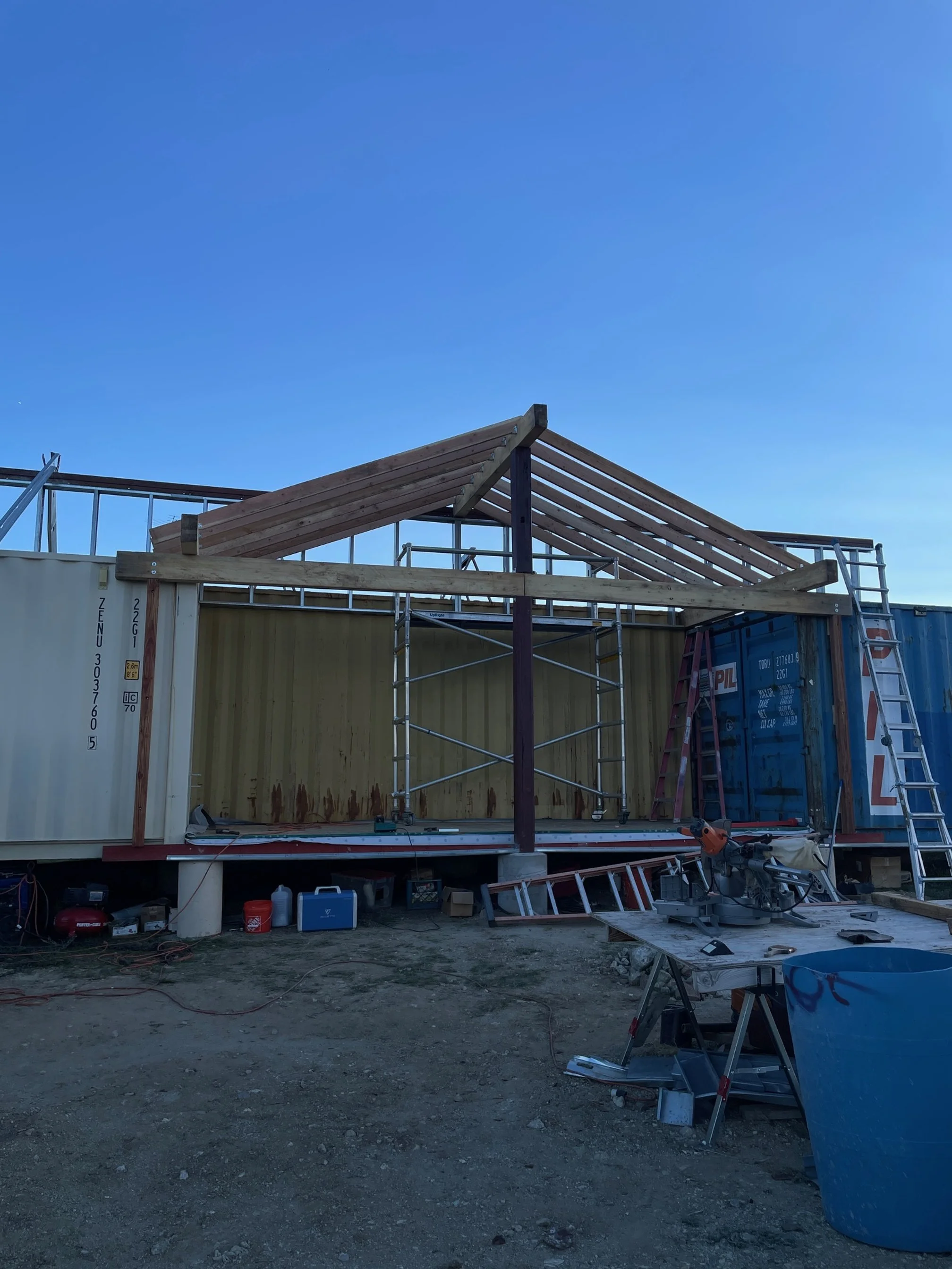 Shipping container home build site featuring a partially built wooden roof structure on top of a shipping container, with scaffolding, ladders, and construction tools scattered around under a clear blue sky.
