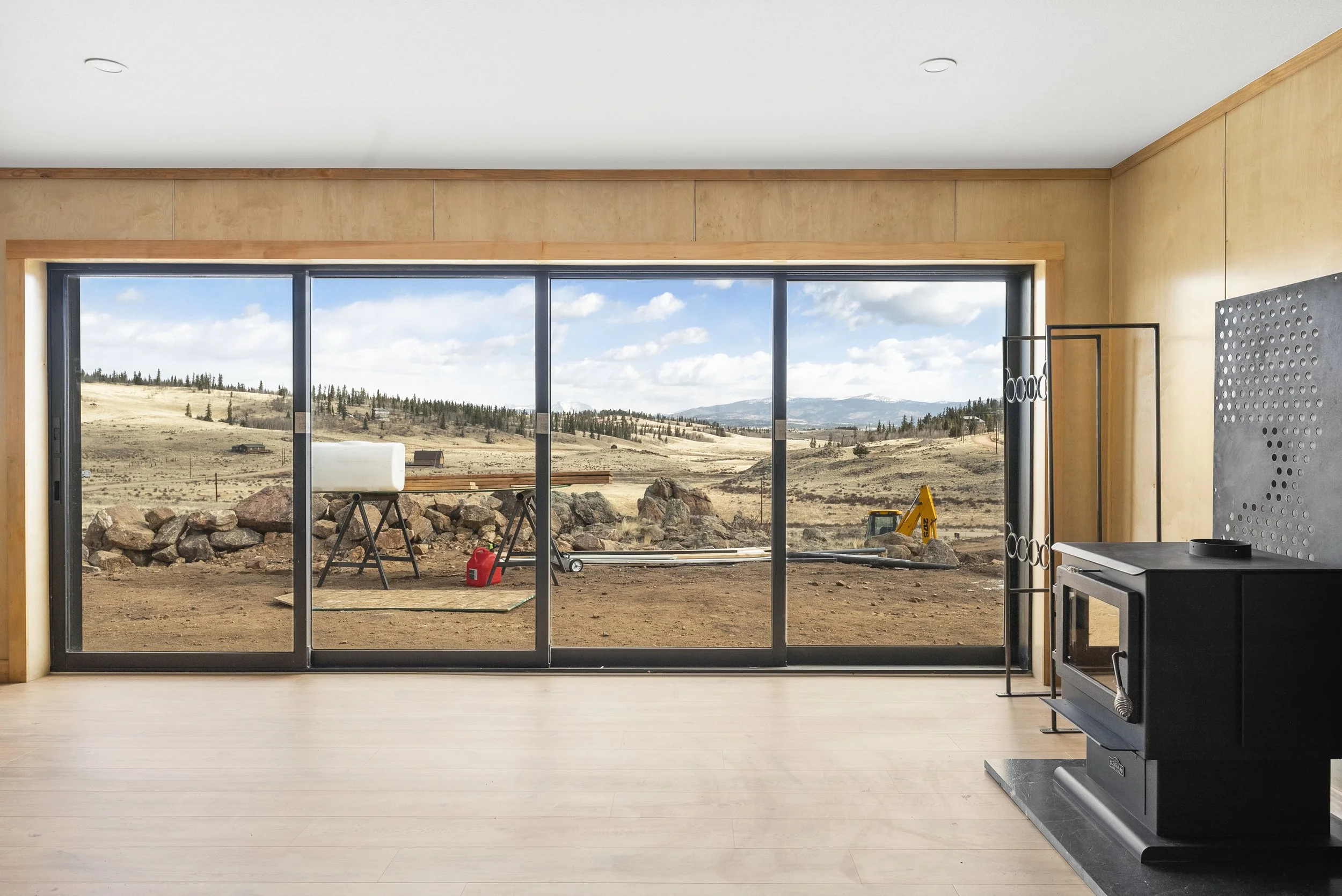 Empty living room with a large glass sliding door overlooking a construction site and a rural landscape with rolling hills, scattered trees, and a partly cloudy sky.