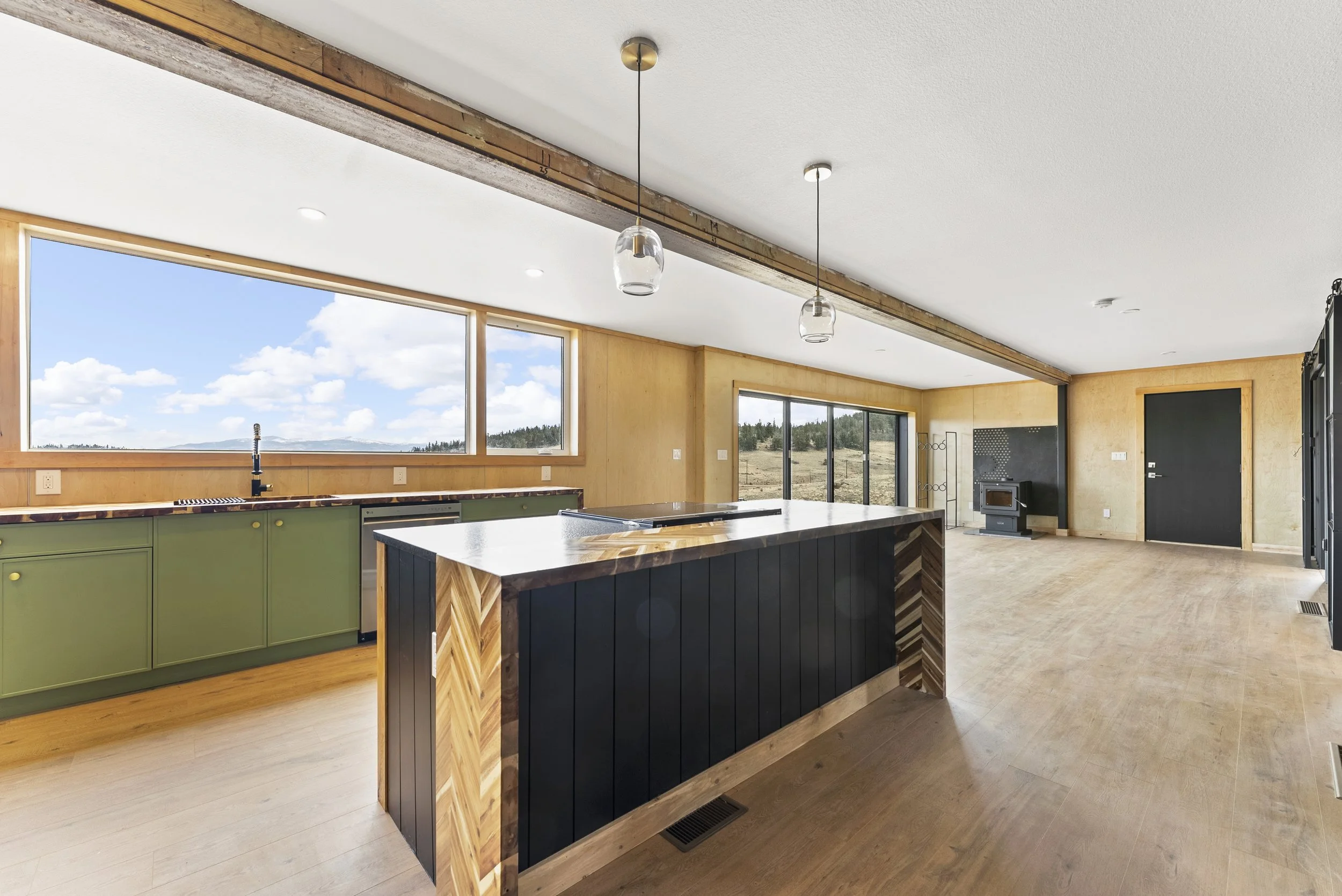Open concept kitchen and living space with large windows, green lower kitchen cabinets, a black kitchen island with wooden accents, and a wood stove in the distance.