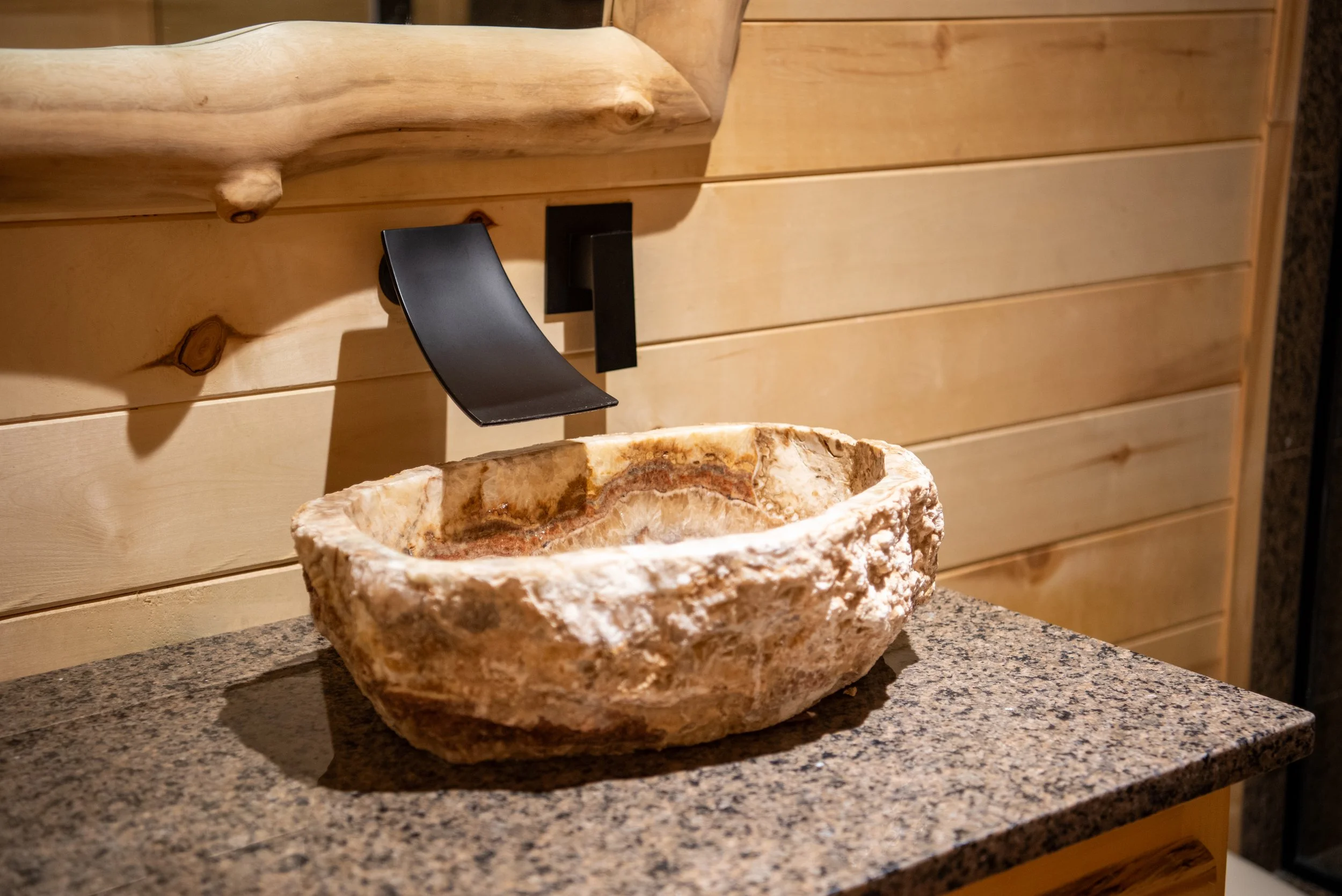 Natural stone sink on granite countertop in a bathroom with wood paneling and a black faucet.