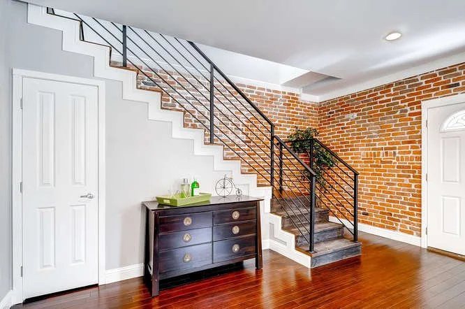Interior of a home featuring a staircase with black metal railing, brick wall background, white door, and dark wood flooring.