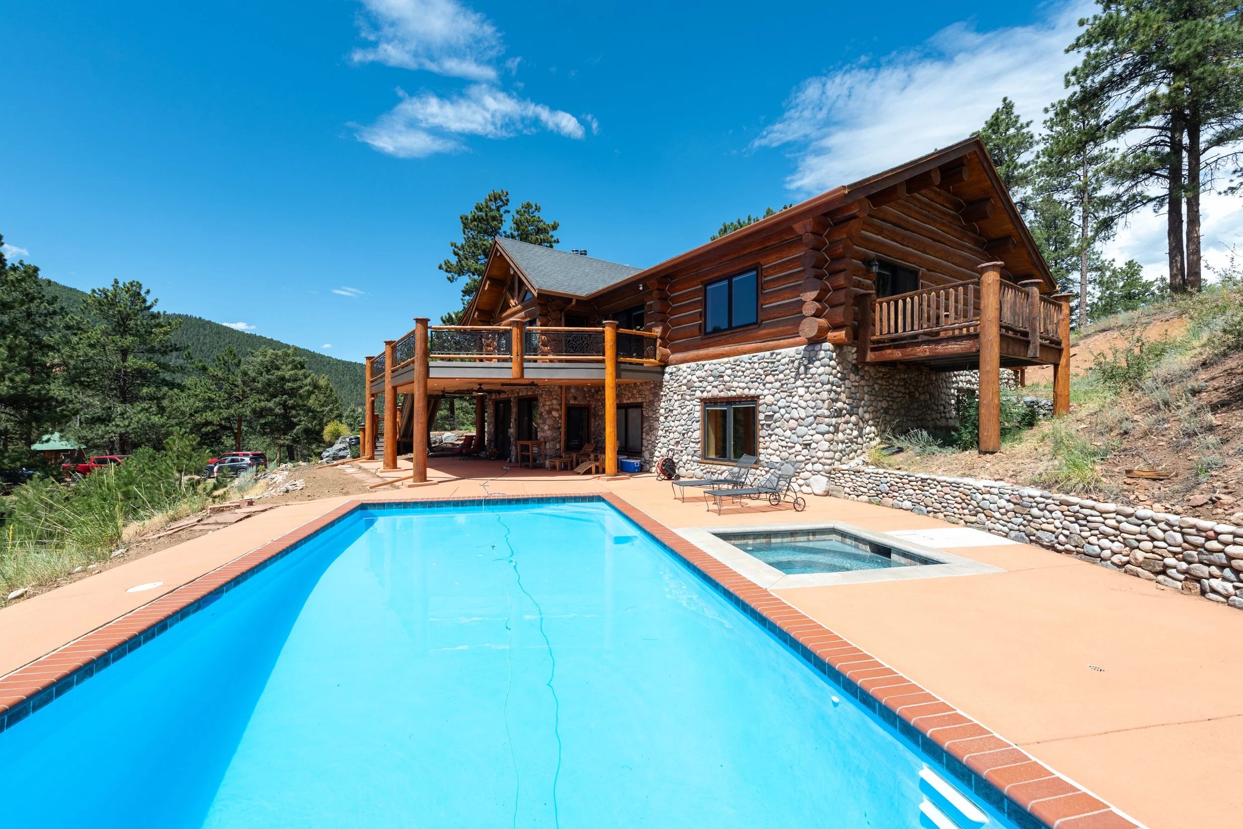 A large wooden and stone mountain house with a deck, outdoor chairs, and a swimming pool with a hot tub in the backyard. Situated in a wooded area under a blue sky with clouds.