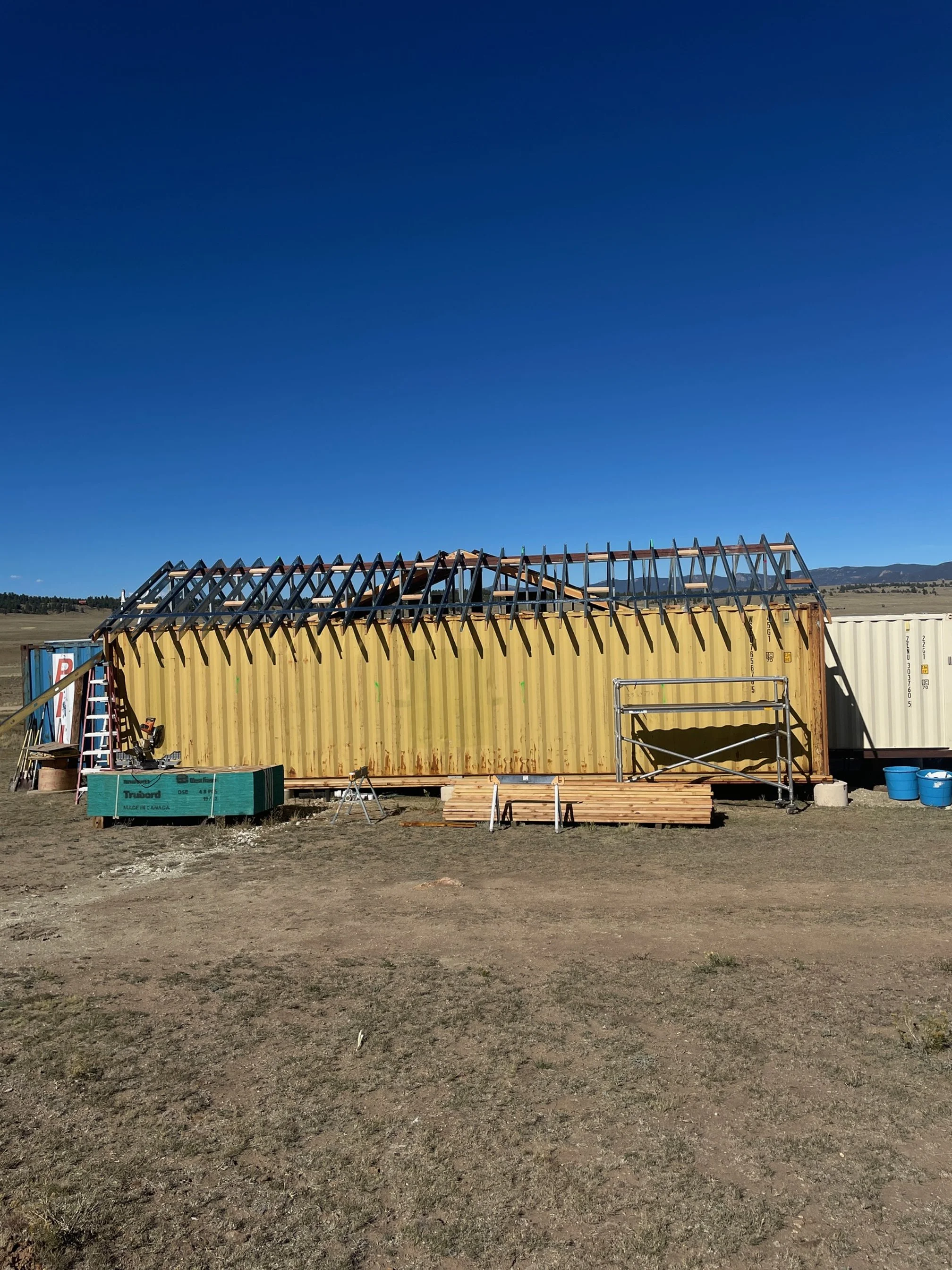 shipping container home build in progress, scaffolding, tools, and building materials scattered around, set on a dirt ground under a clear blue sky.