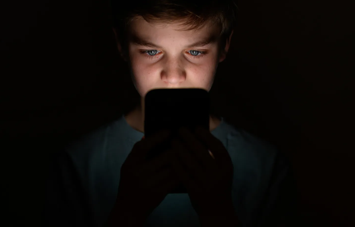 A young boy with blue eyes looking at a smartphone in a dark room.