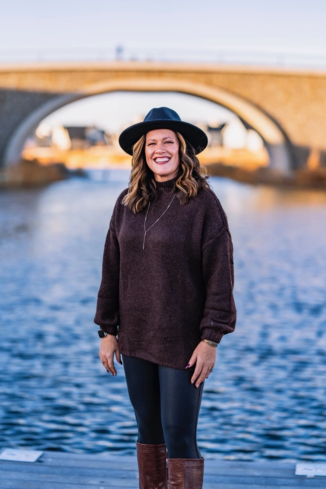 A woman standing outdoors near a body of water, smiling, wearing a black hat, dark sweater, black pants, and brown boots, with a bridge in the background.