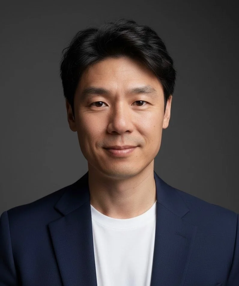 Headshot of a man with black hair wearing a navy blazer and white t-shirt against a gray background.