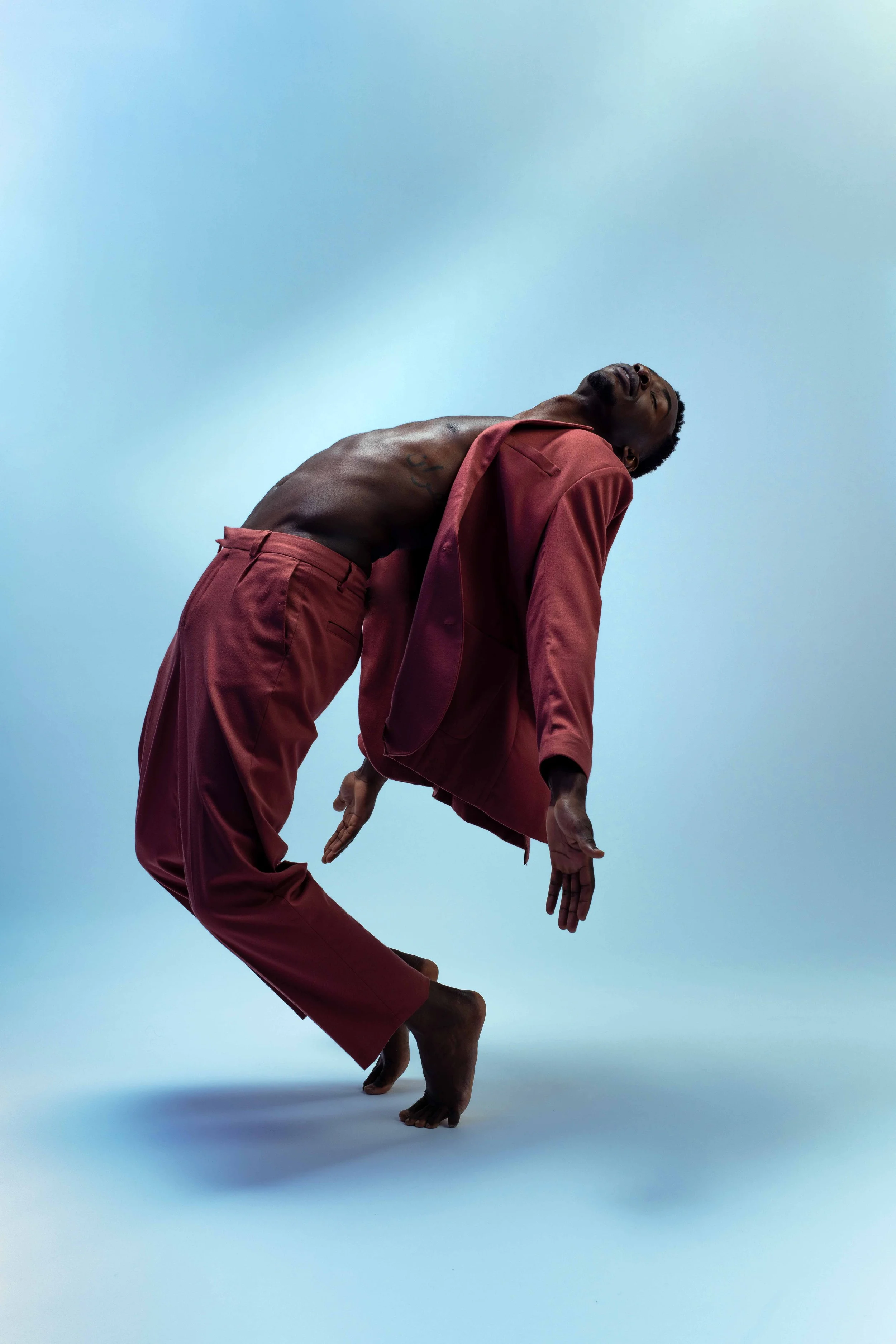 A man with dark skin, shirtless except for maroon pants, is bent backward with his head tilted back and arms loose, wearing a maroon blazer draped over his shoulders, against a light blue background.
