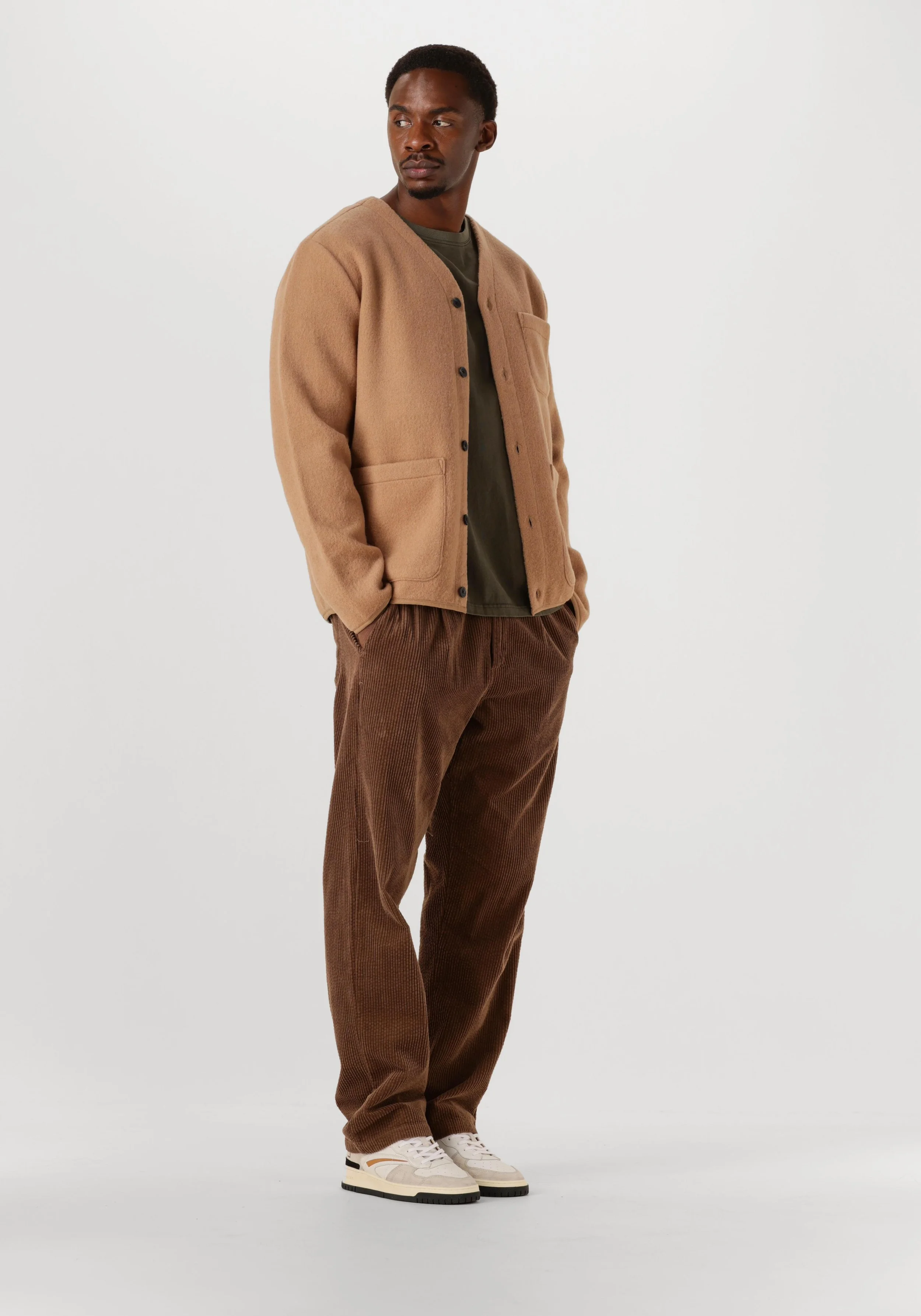 A man standing against a plain white background wearing a tan cardigan, dark green t-shirt, brown corduroy pants, and white sneakers.