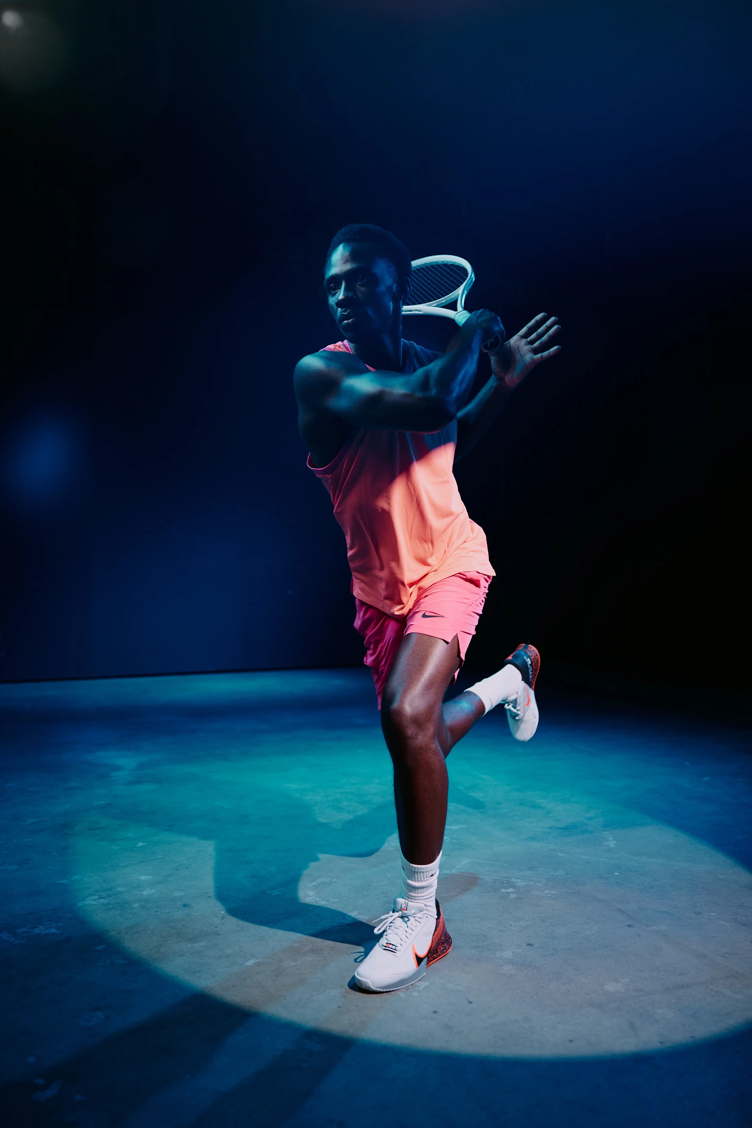 A male tennis player in athletic gear swings a tennis racket inside a dark, stage-like setting under spotlight.