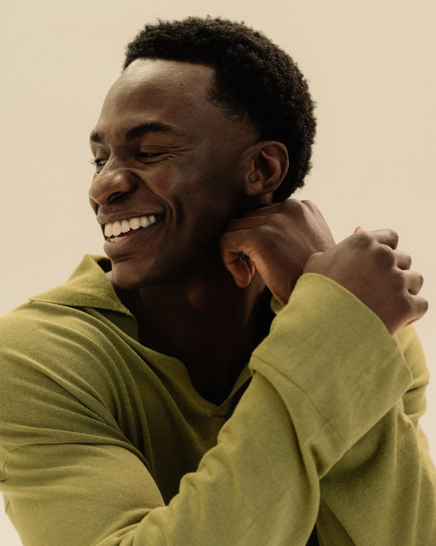 A smiling man with short curly hair, wearing a yellow hoodie, poses with his head turned to the side and his left hand resting near his face.