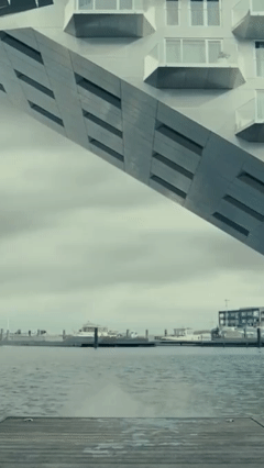 Modern building with unique angled design and overhanging sections seen from across a body of water with cloudy sky in background.