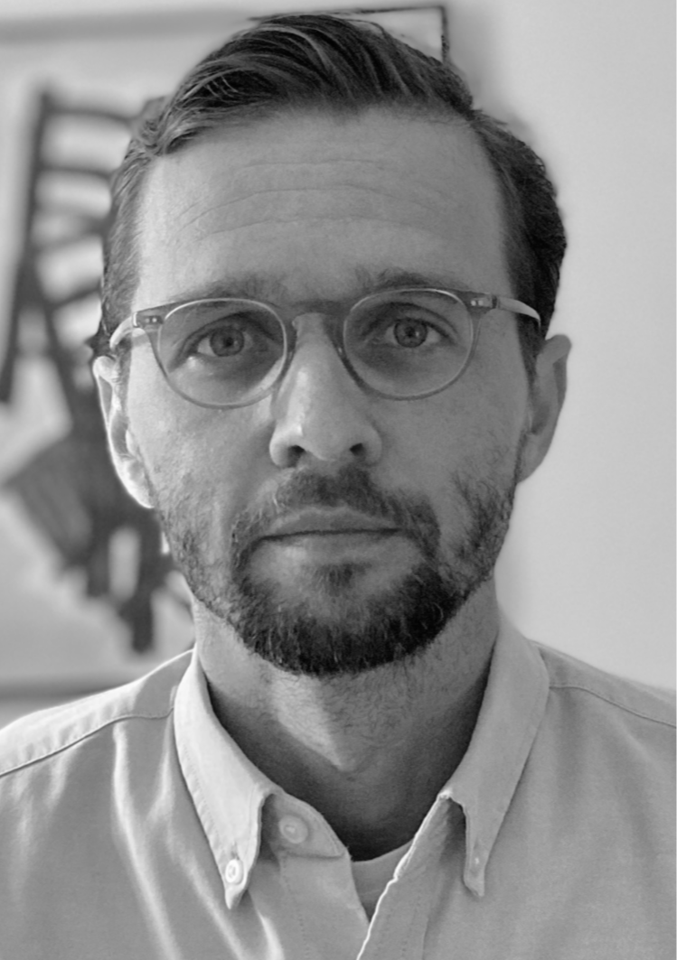 Homem de óculos de armação fina, barba por fazer, cabelo penteado para trás, usando camisa de botão clara, olhando diretamente para a câmera em fundo desfocado.