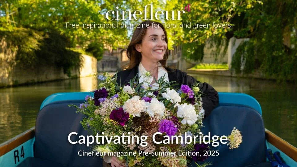 A woman enjoying a boat ride on a river during the daytime with lush green trees surrounding the water. She is holding a large bouquet of mixed flowers and smiling.