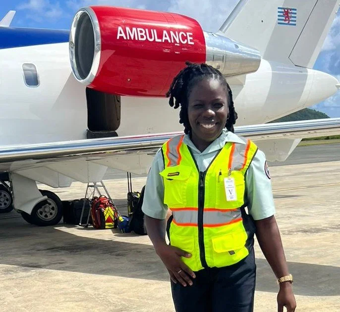 Female emergency medical technician in uniform and yellow safety vest standing in front of an ambulance jet on an airfield.