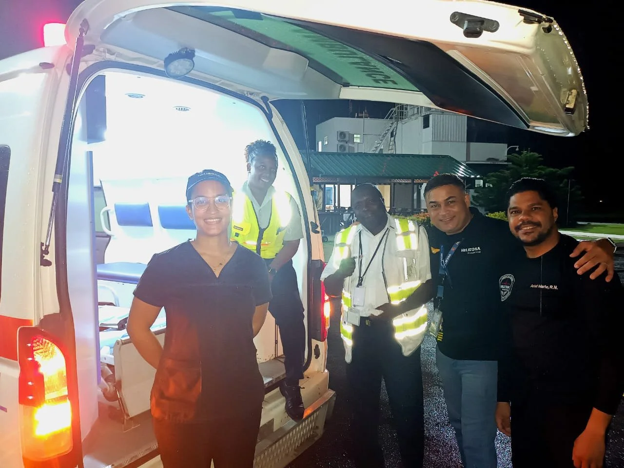 Five emergency responders standing next to an ambulance at night. One is inside the ambulance, and four are outside, smiling at the camera. They are wearing uniforms, with some in reflective vests.