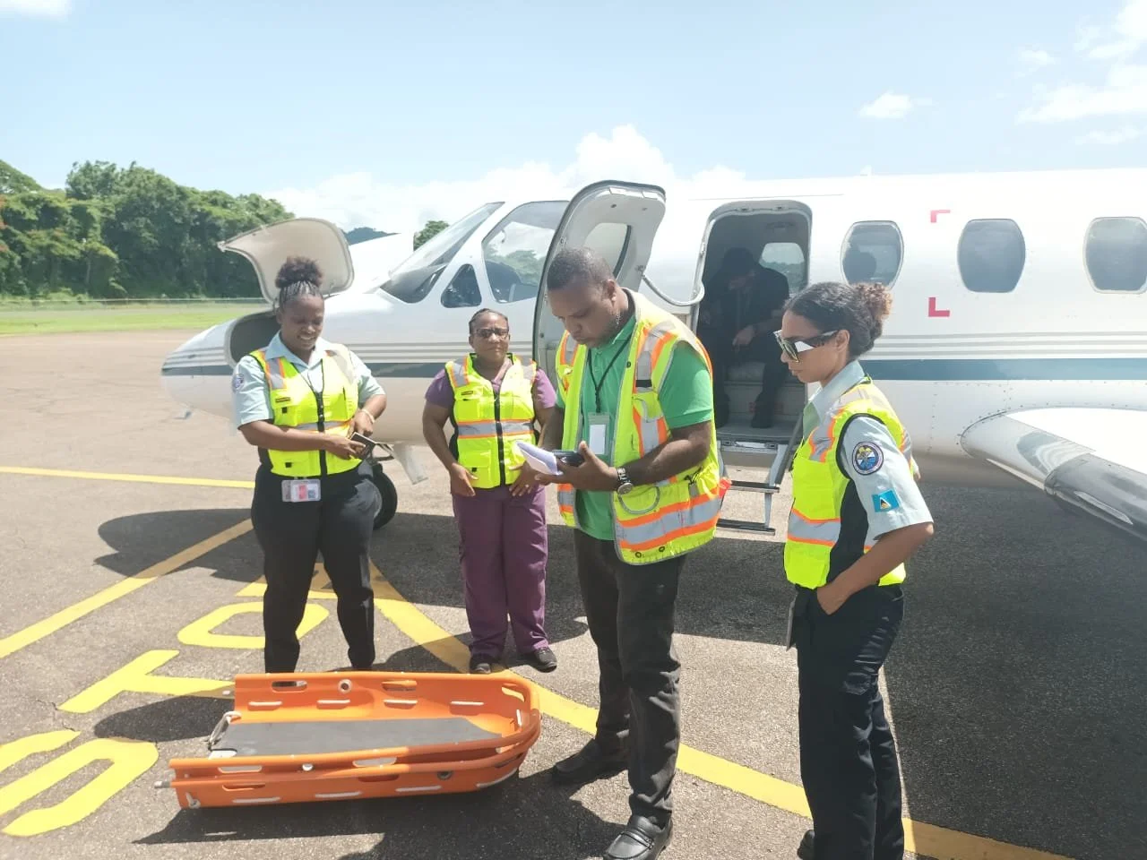Group of airport workers standing near a private jet, one person is writing on a notepad, others are observing, with a stretcher on the ground.