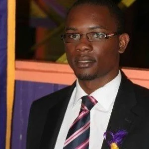 A man in a suit with glasses and a striped tie, standing outdoors with colorful background.