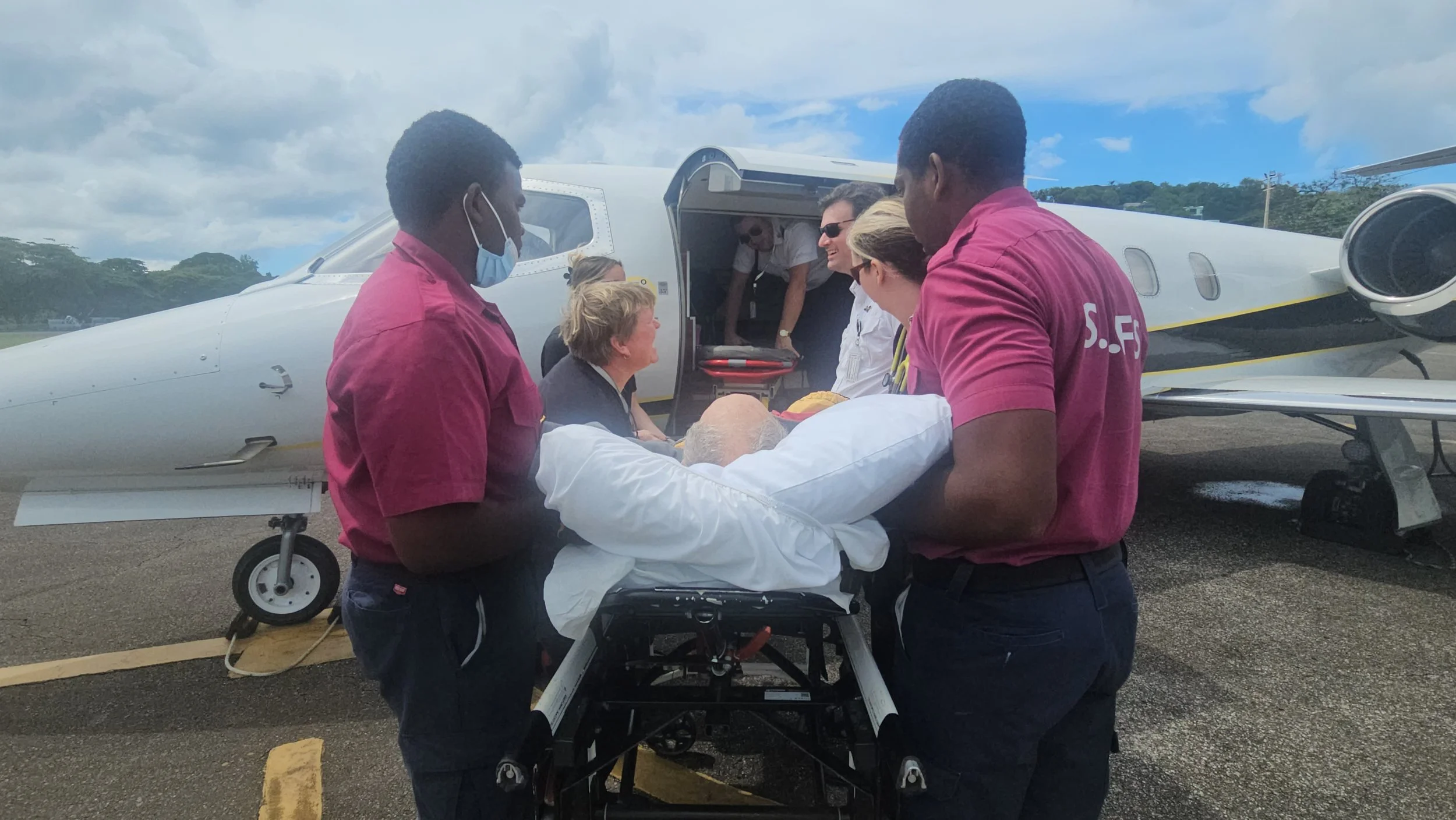 Medical team transporting a patient on a stretcher from a private jet, with medical staff assisting and the patient covered with a white blanket, in an outdoor airport setting.