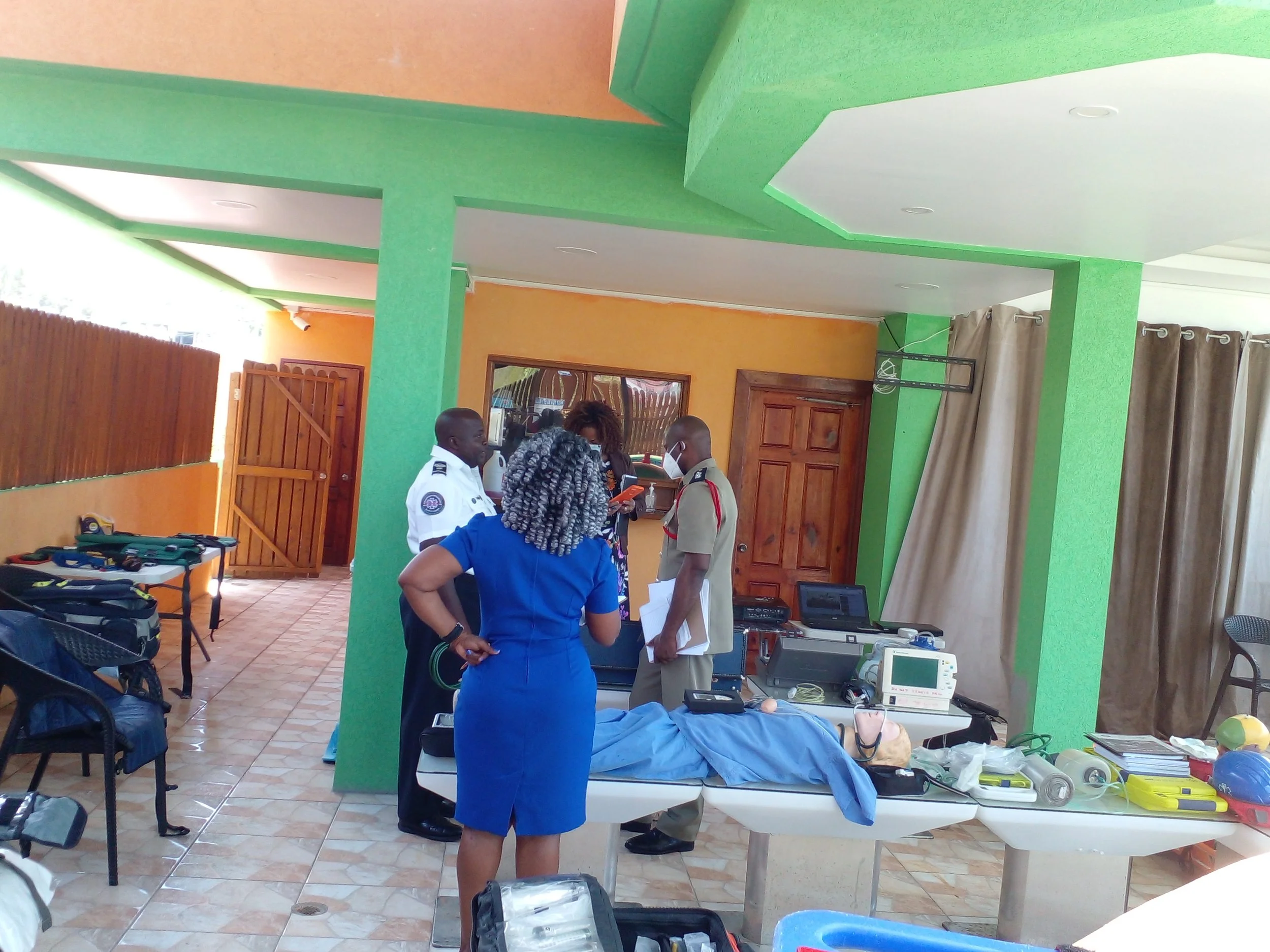 Medical personnel and officials gather around a table with medical supplies and equipment in a brightly colored room with green and orange walls. There are chairs, curtains, and a wooden door in the background.