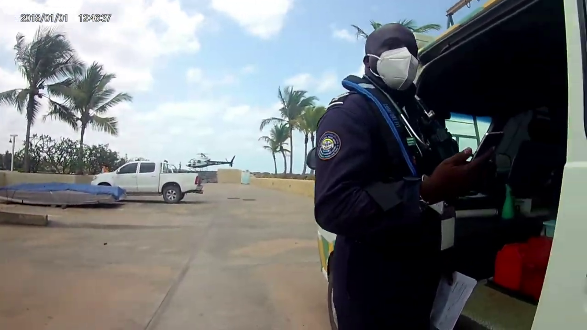 A man wearing a face mask and a jacket with an insignia on the sleeve, standing next to a vehicle, looking at a device, with palm trees and a helicopter in the background.