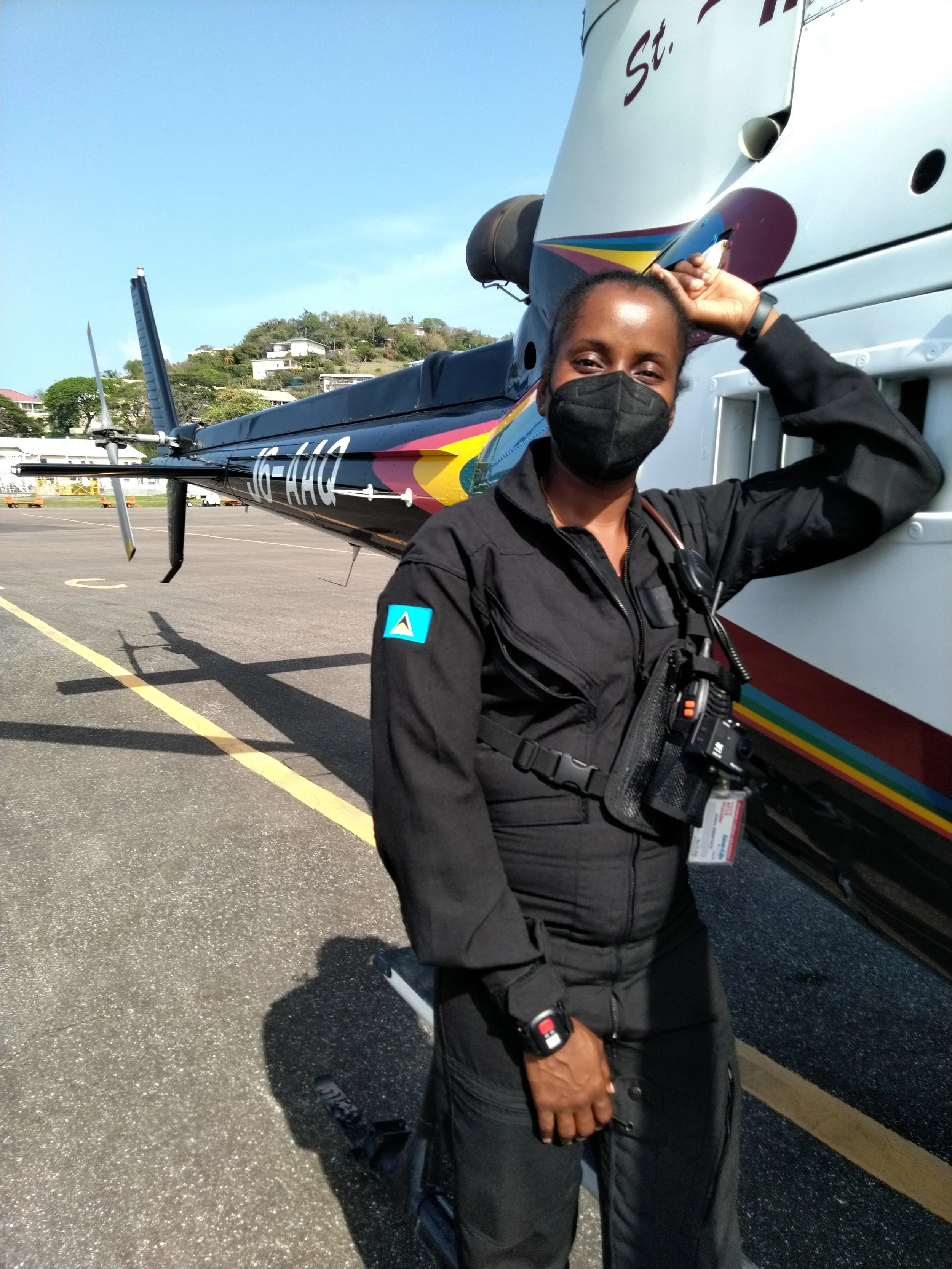 A woman dressed in black aviation uniform, wearing a black face mask, standing next to a helicopter, holding her head with her right hand, with a hillside and residential buildings in the background.