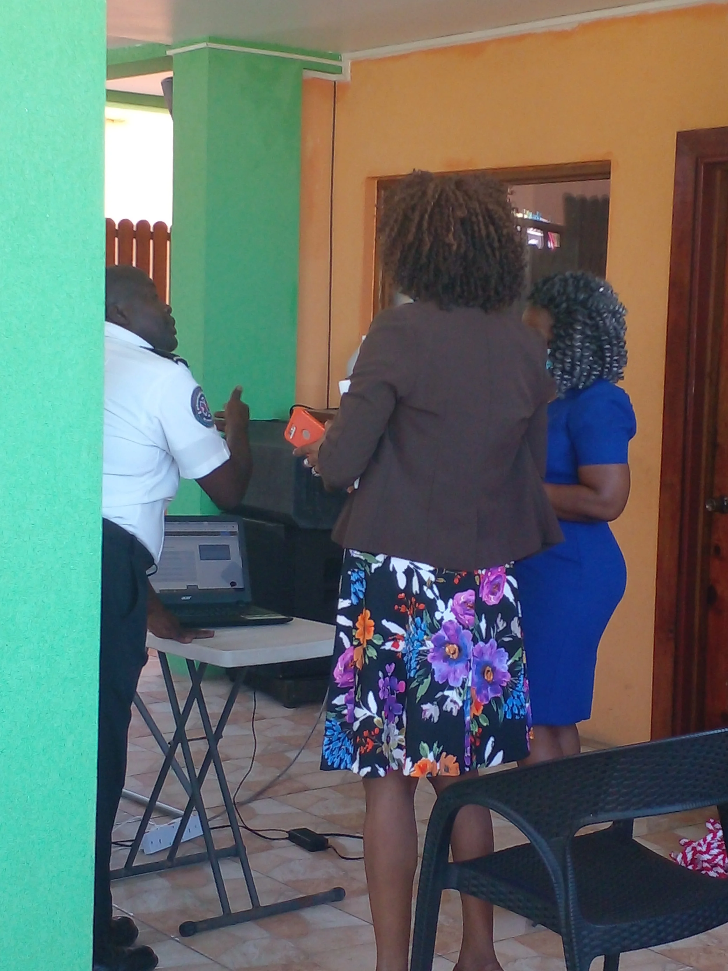 Three women and a man gathered around a laptop and printer in a room with tan and green walls, engaged in a discussion.
