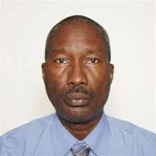 A middle-aged African American man with a serious expression, wearing a light blue shirt and a patterned tie, against a plain white background.