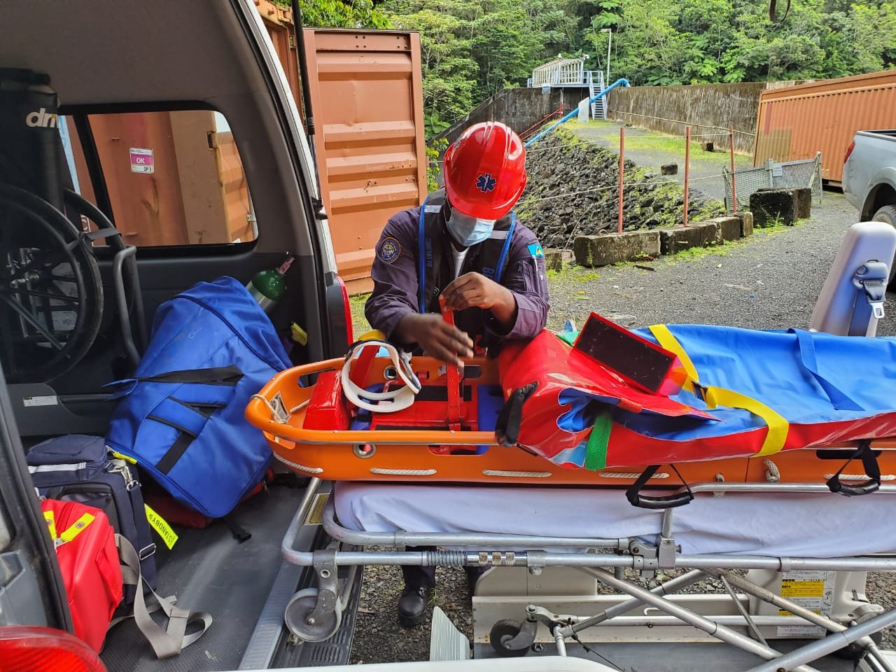 Paramedic wearing a red helmet and face mask preparing a stretcher with medical equipment outdoors near an ambulance.