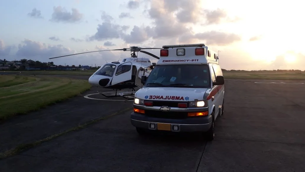 An ambulance and a helicopter on a tarmac at sunset with a cloudy sky.
