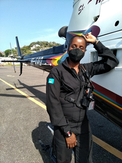 A woman in a black uniform and face mask standing next to a helicopter on a tarmac, with her hand raised and holding a helmet.