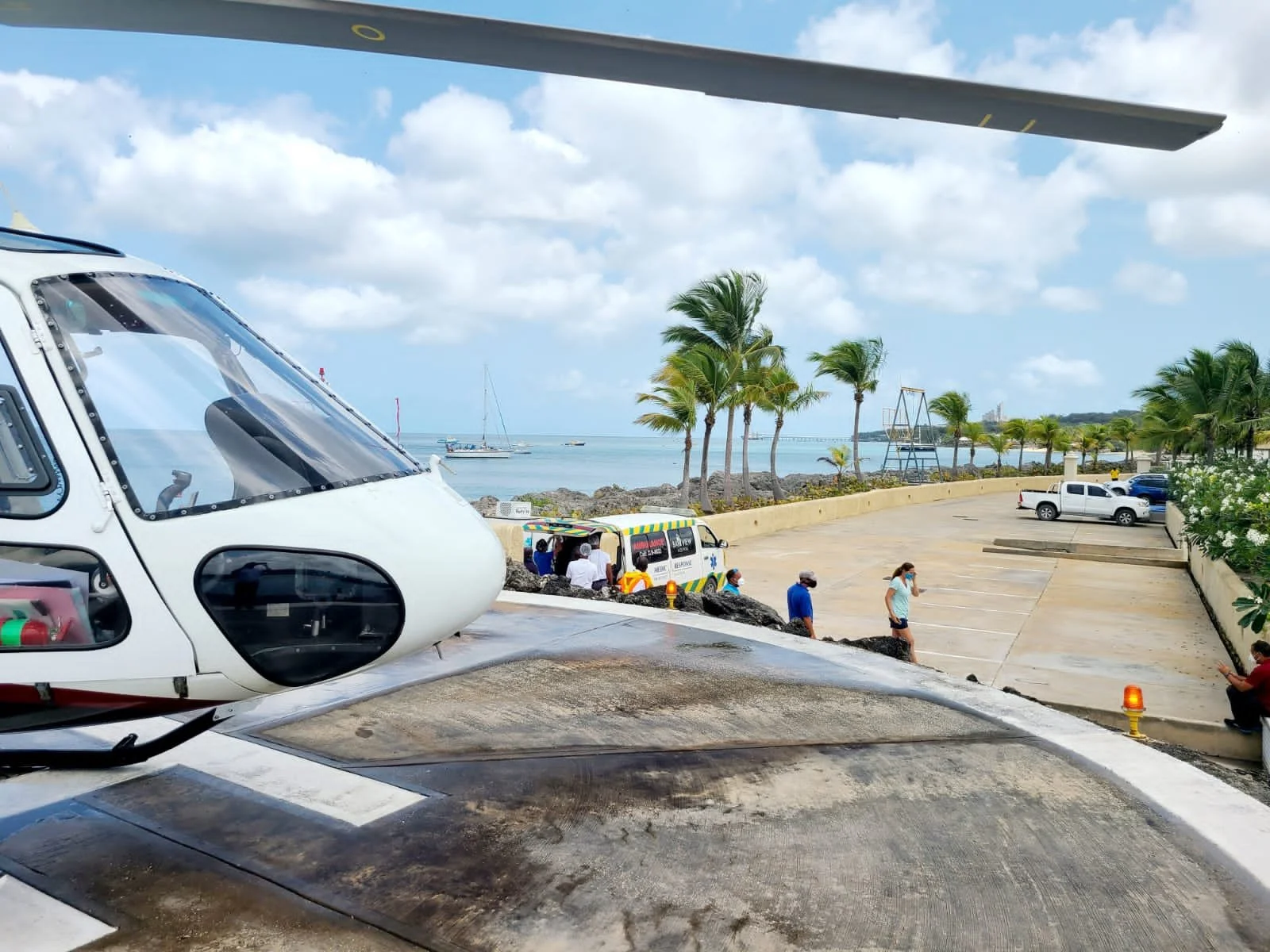 A helicopter in the foreground with a white body and black and red accents, parked near a coastal area with palm trees, boats in the water, and people walking near a parking lot.