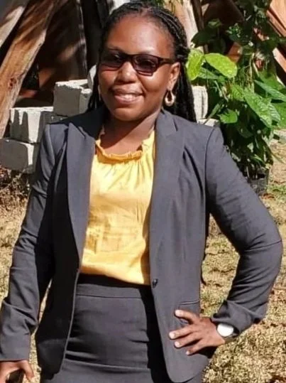 A woman in business attire, wearing sunglasses and earrings, standing outdoors with her left hand on her hip and holding a phone in her right hand, in front of a rustic background with wooden structures and greenery.