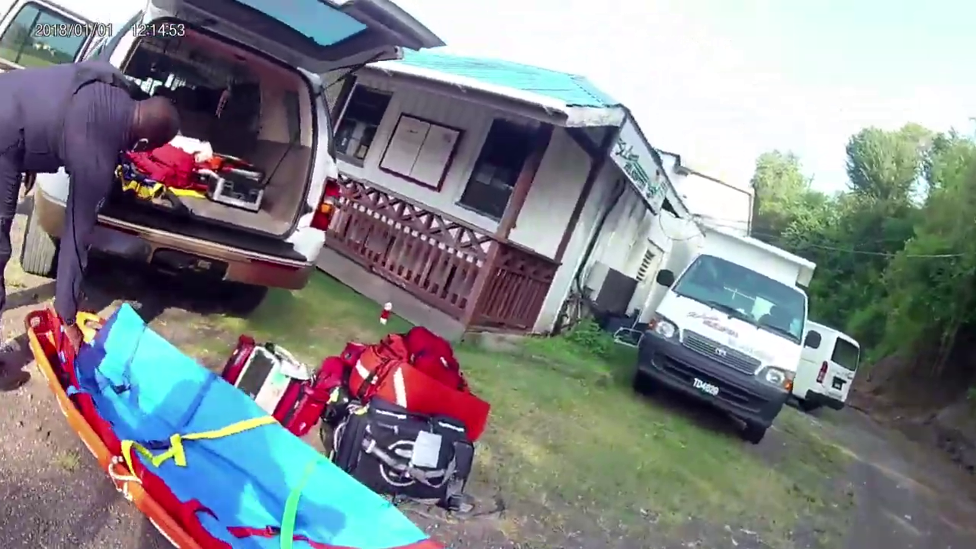 Emergency medical personnel preparing rescue equipment outside a mobile home, with rescue gear and a backboard on the ground nearby. A man is reaching into an open ambulance trunk. Two white vans are parked in front of the mobile home, which is surro