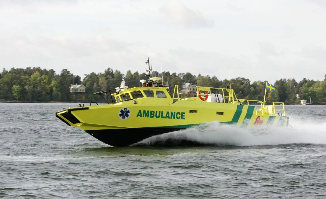 A yellow boat with the word 'AMBULANCE' and a medical symbol on the side, speeding across a body of water, with a Swedish flag at the back and a city skyline with trees in the background.