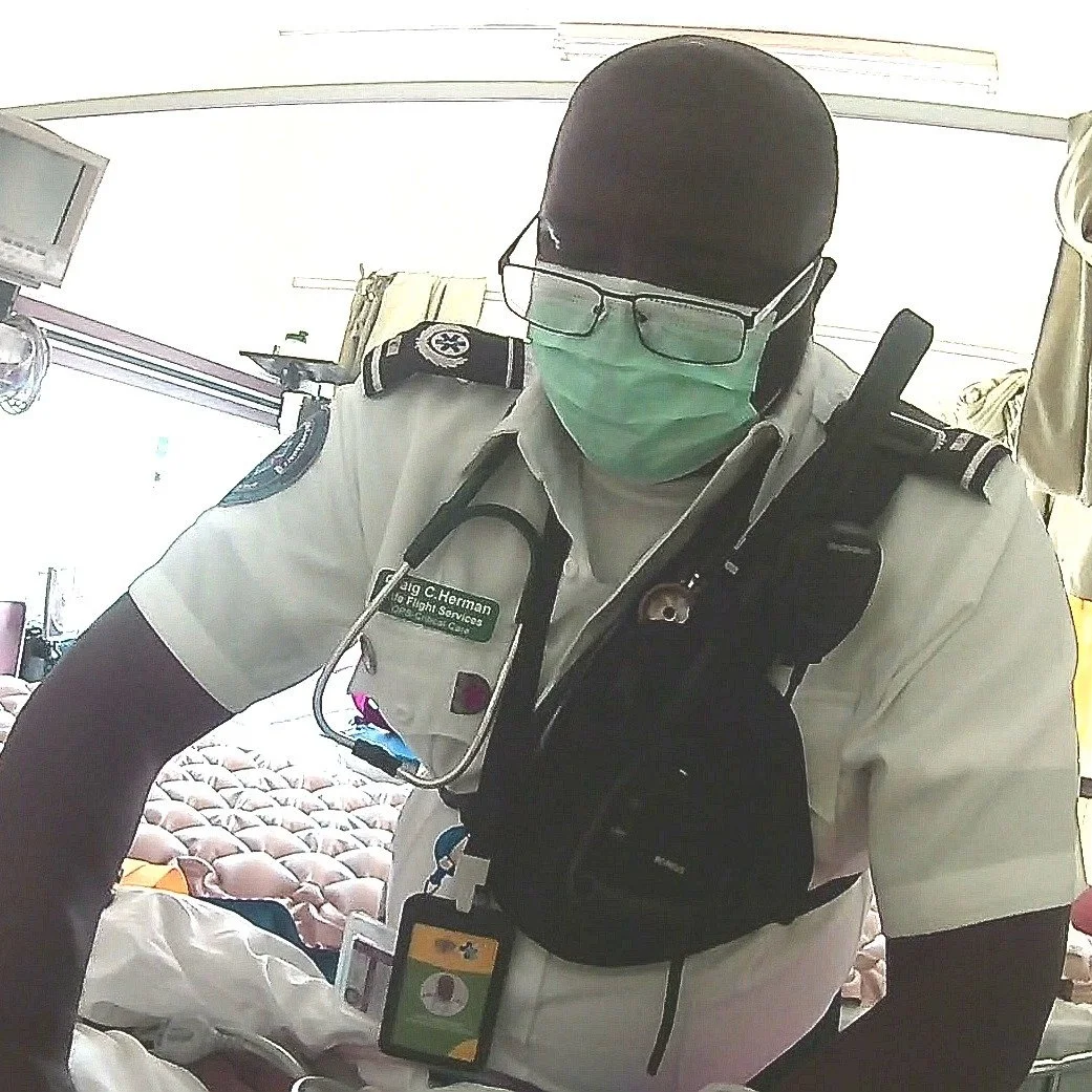A paramedic in uniform with a face mask, glasses, and a stethoscope around his neck, taking a selfie inside a medical facility.
