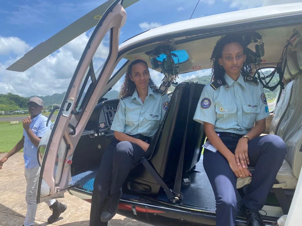 Two female pilots sitting inside a helicopter while a man walks by outside, with a cloudy blue sky in the background.