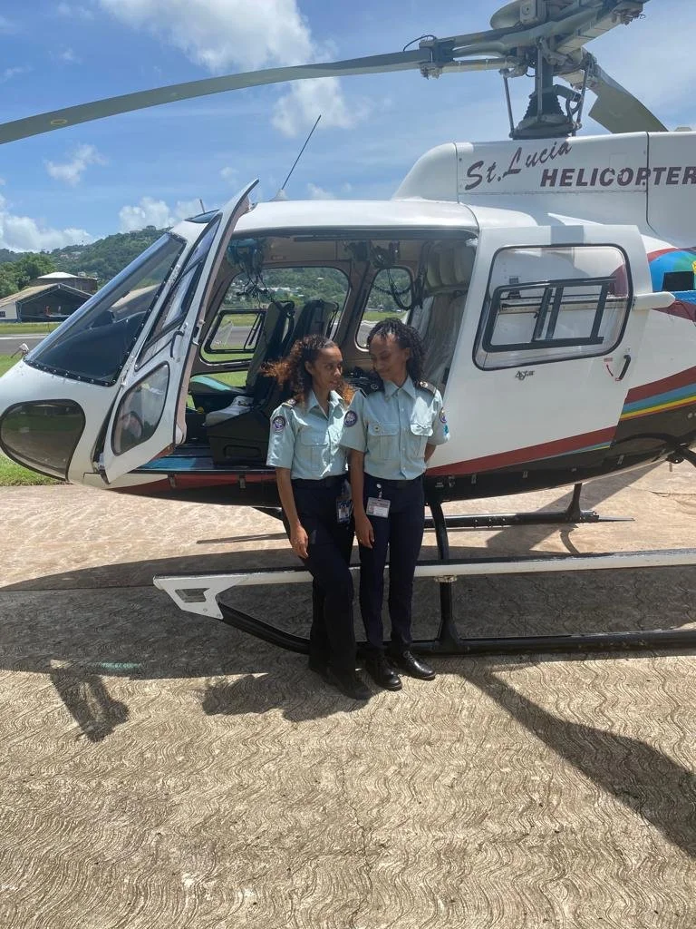 Two women in pilot uniforms standing in front of a helicopter on a tarmac. The helicopter is white with markings and the door open, revealing the interior. The background shows a grassy area, some trees, and a partly cloudy sky.