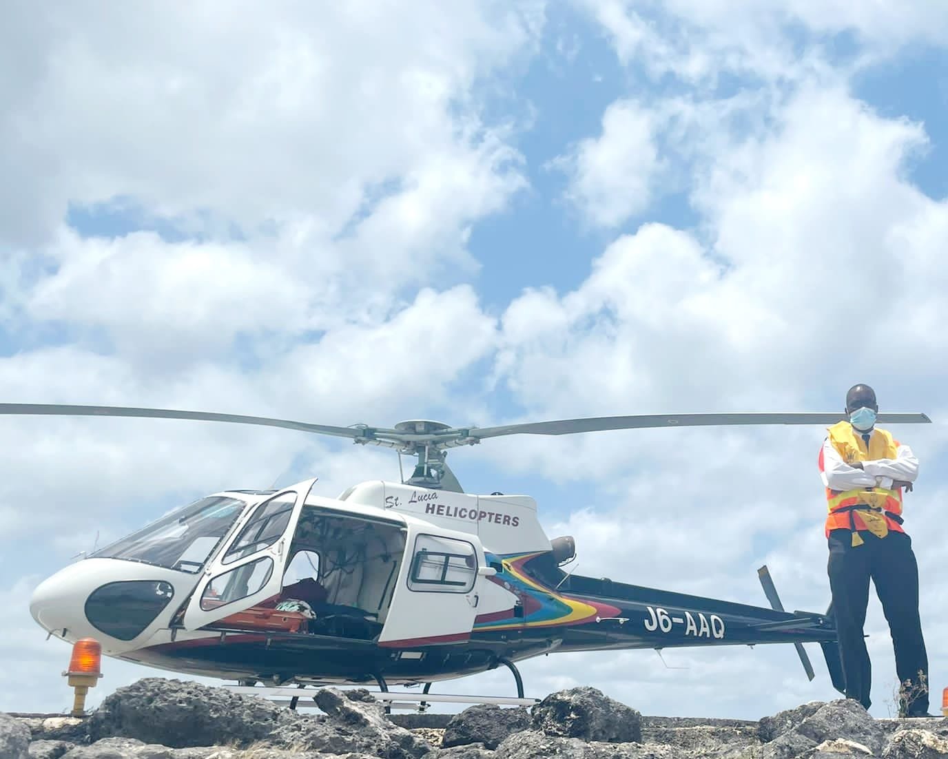 A helicopter on rocky terrain with a person standing nearby wearing a yellow safety vest, face mask, sunglasses, and a helmet under a cloudy sky.
