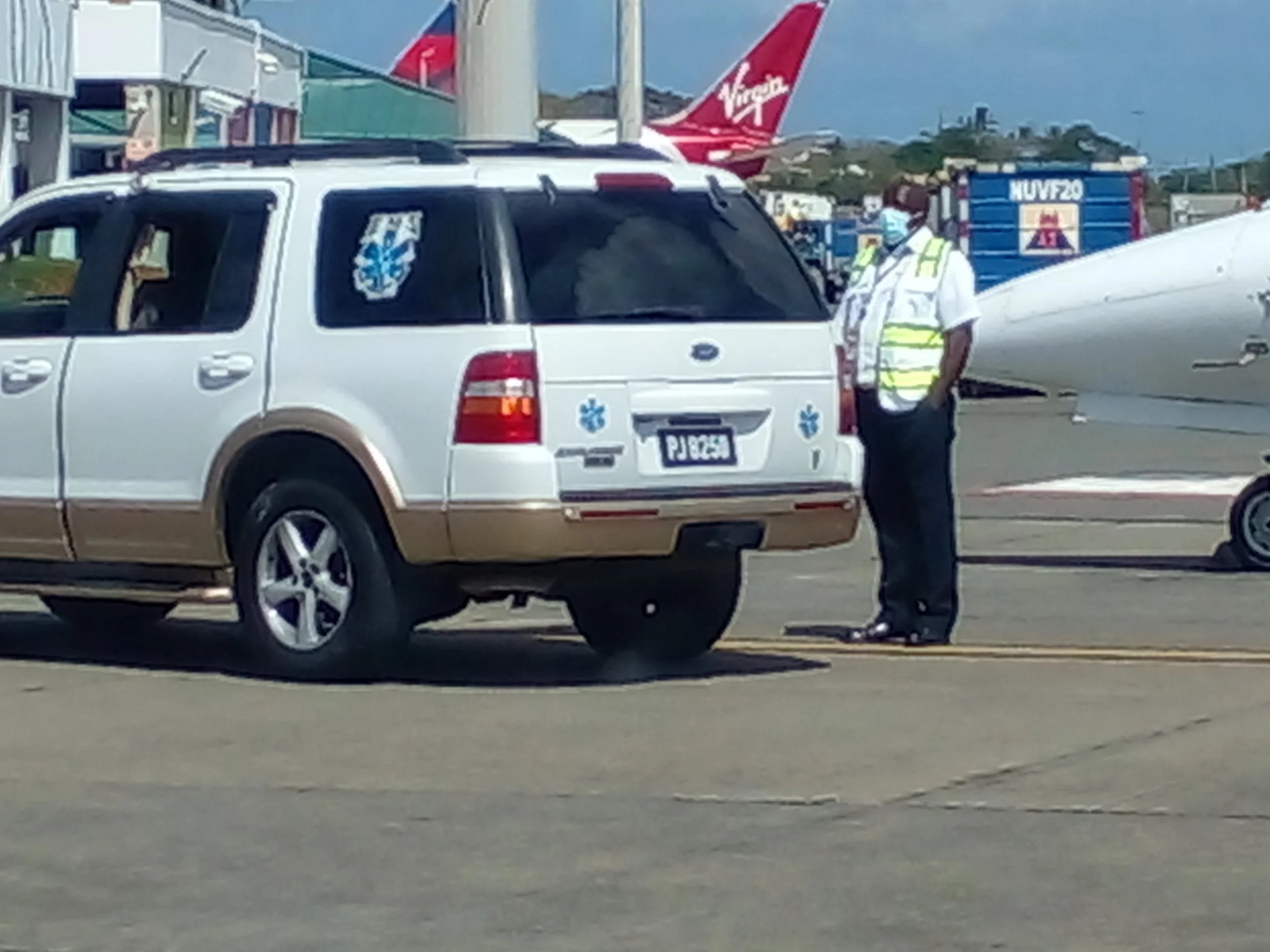 A man in a white shirt, reflective safety vest, and face mask standing next to a white ambulance vehicle with medical symbols, at an airport tarmac where a Virgin Airlines airplane and other planes are visible in the background.