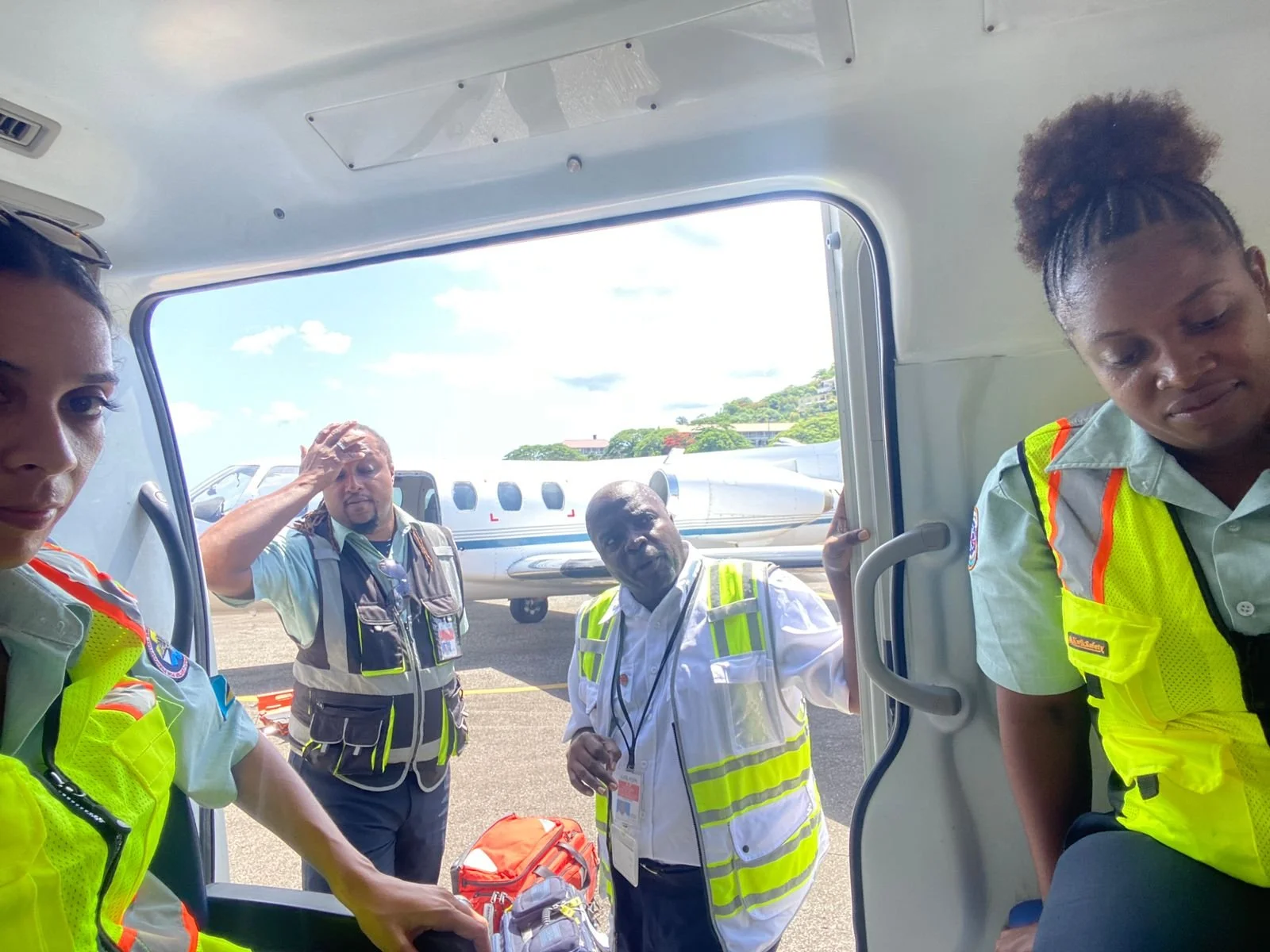 Group of airline crew members and ground staff standing near a small airplane on the tarmac, some wearing high-visibility vests, with a clear sky and green trees in the background.
