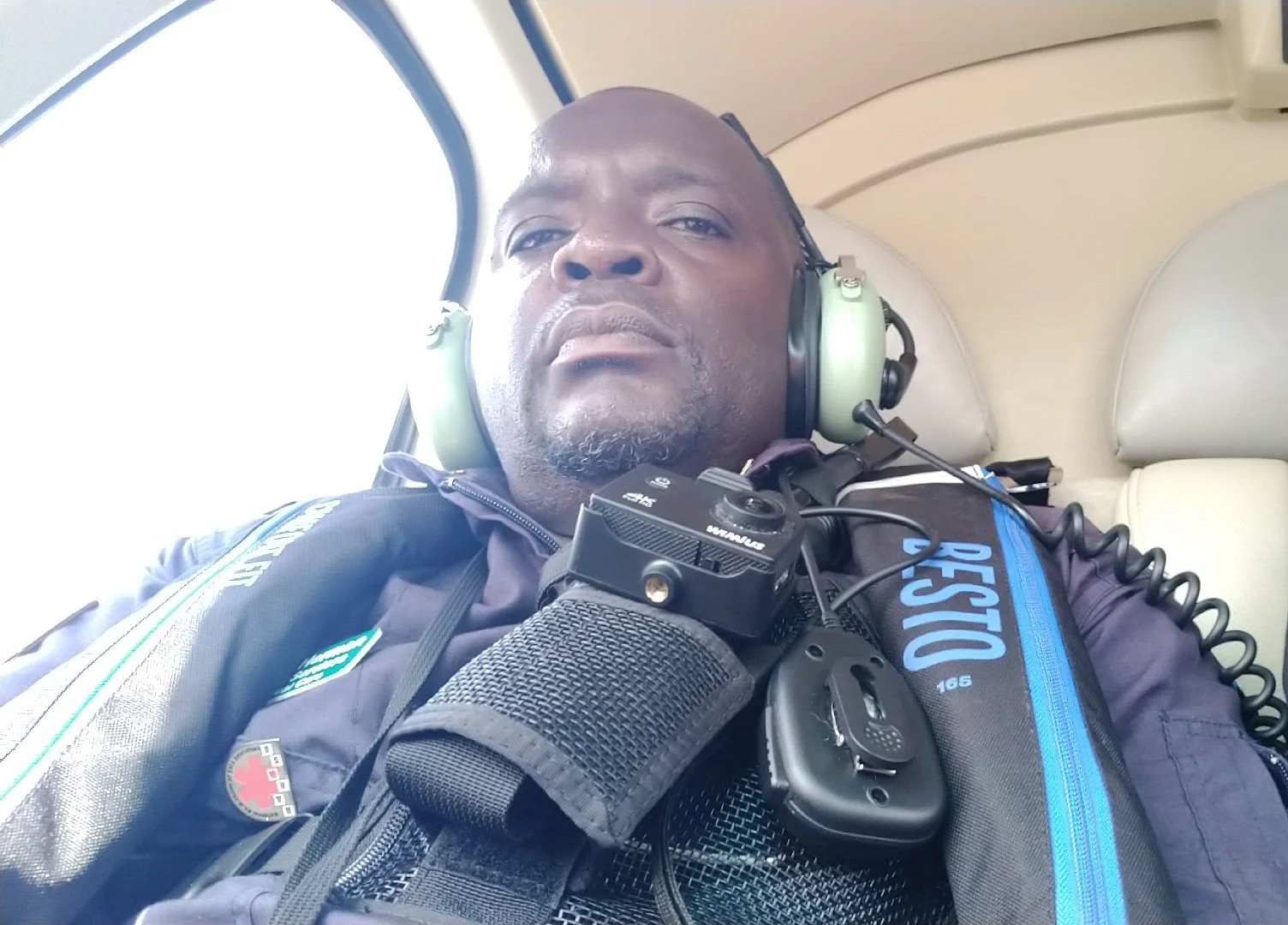 Close-up selfie of a man wearing a headset, vest, and uniform, sitting inside a helicopter or aircraft with a window visible in the background.