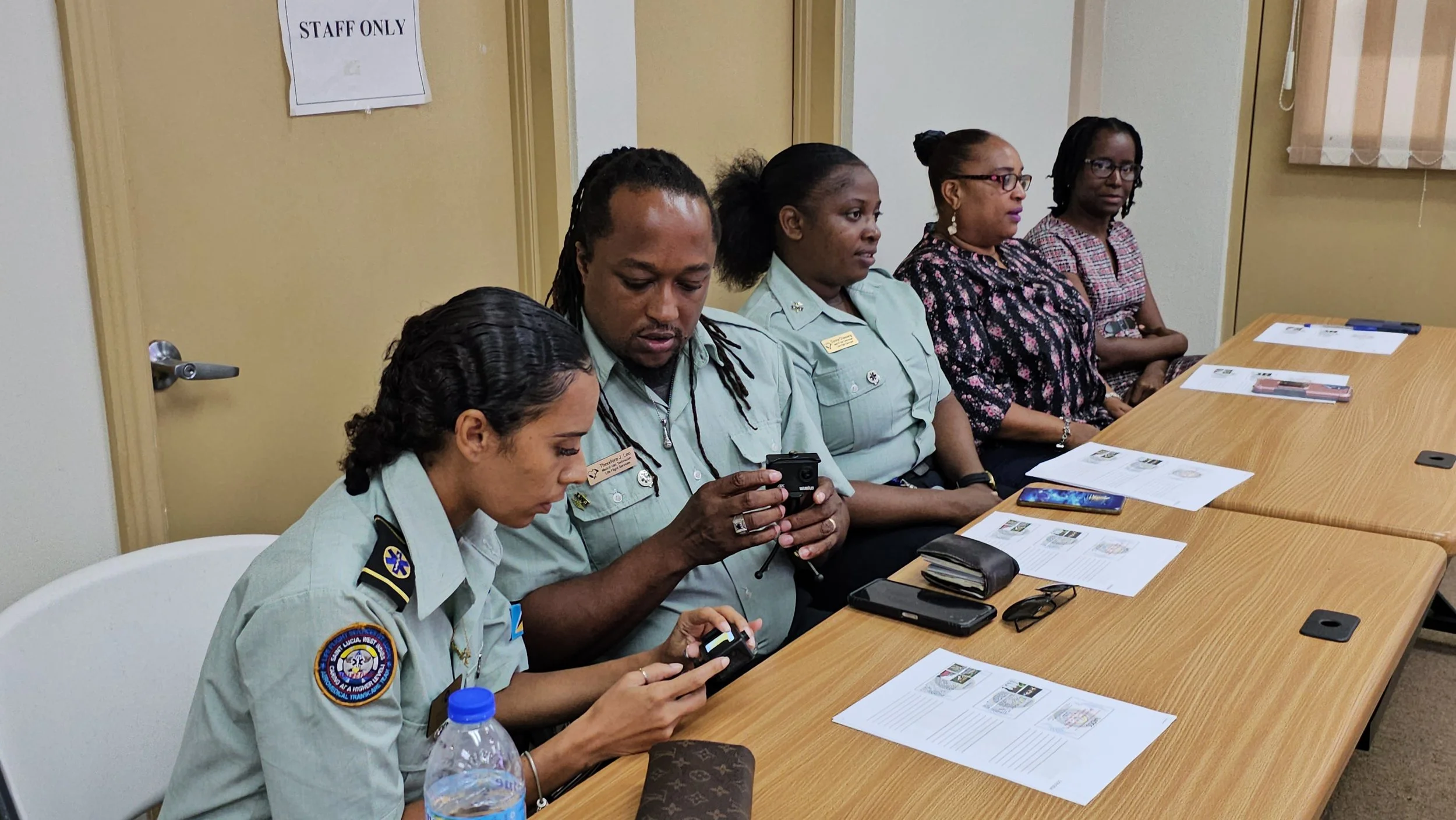 Five women sitting at a conference table, two are in uniform and three are in casual attire, with documents and phones on the table, in a room with a door that says 'STAFF ONLY'.