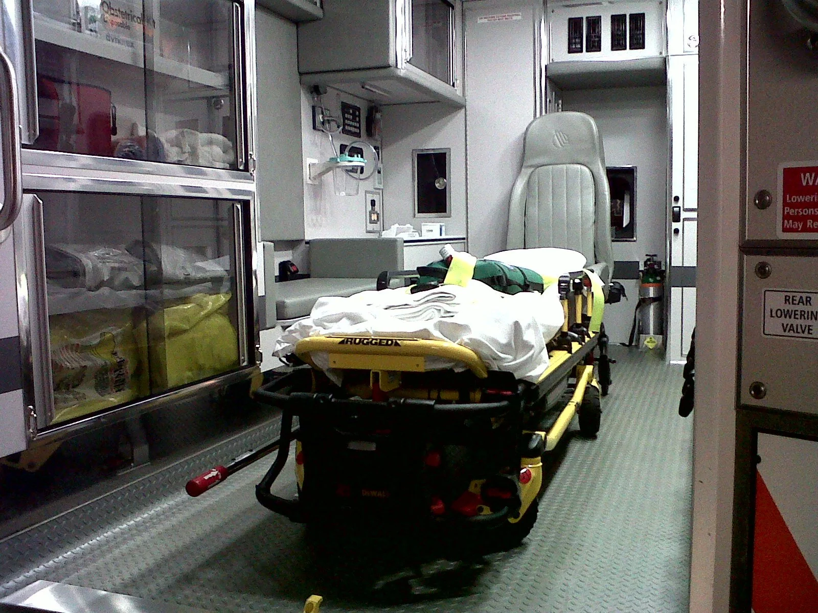 An emergency room in a hospital with a stretcher in the center, covered with white sheets, and medical equipment around. There's an empty medical chair and storage cabinets in the background.