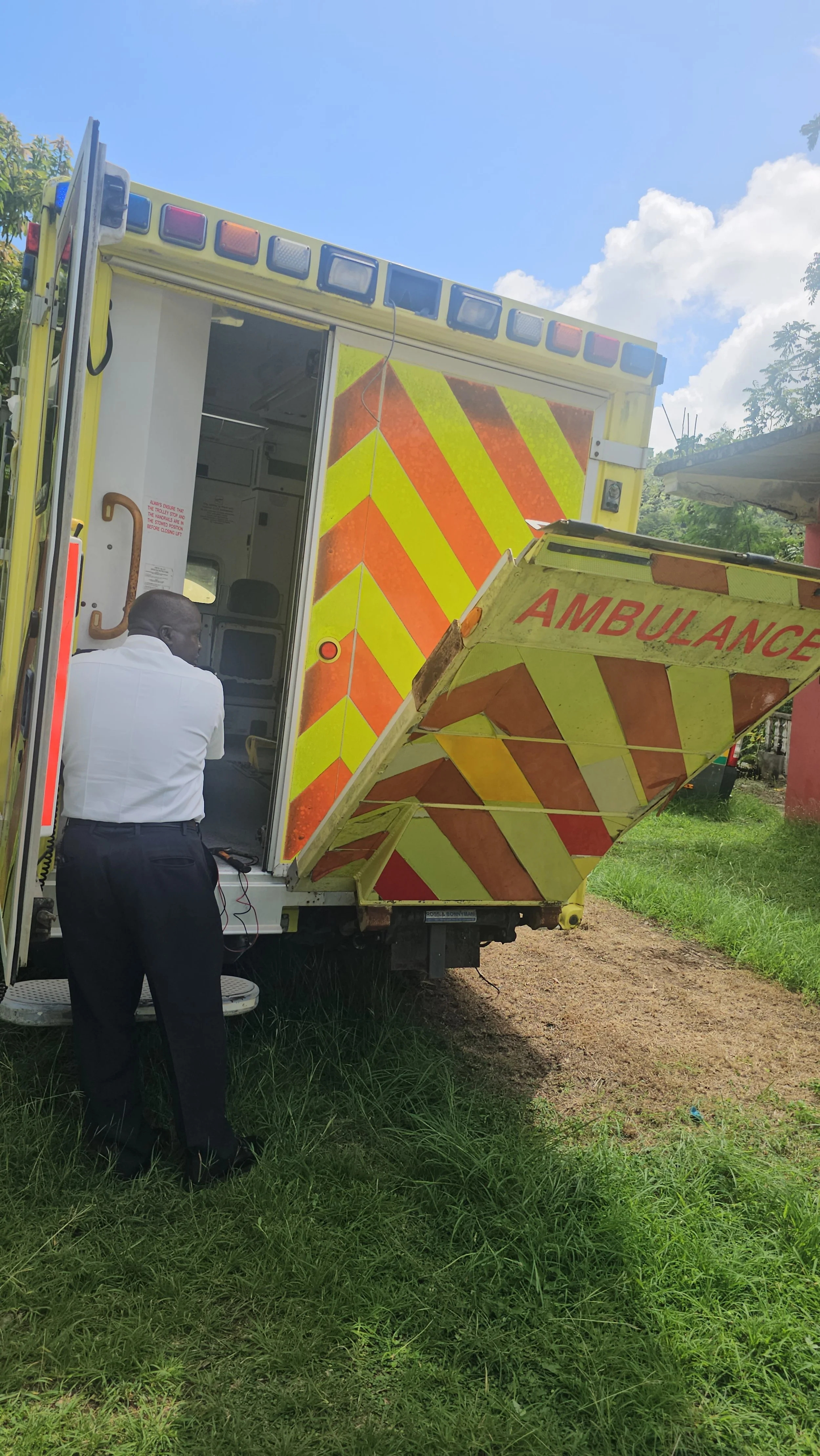 An ambulance parked on a grassy area with a person in white shirt and black pants standing near it, open rear doors showing the interior.