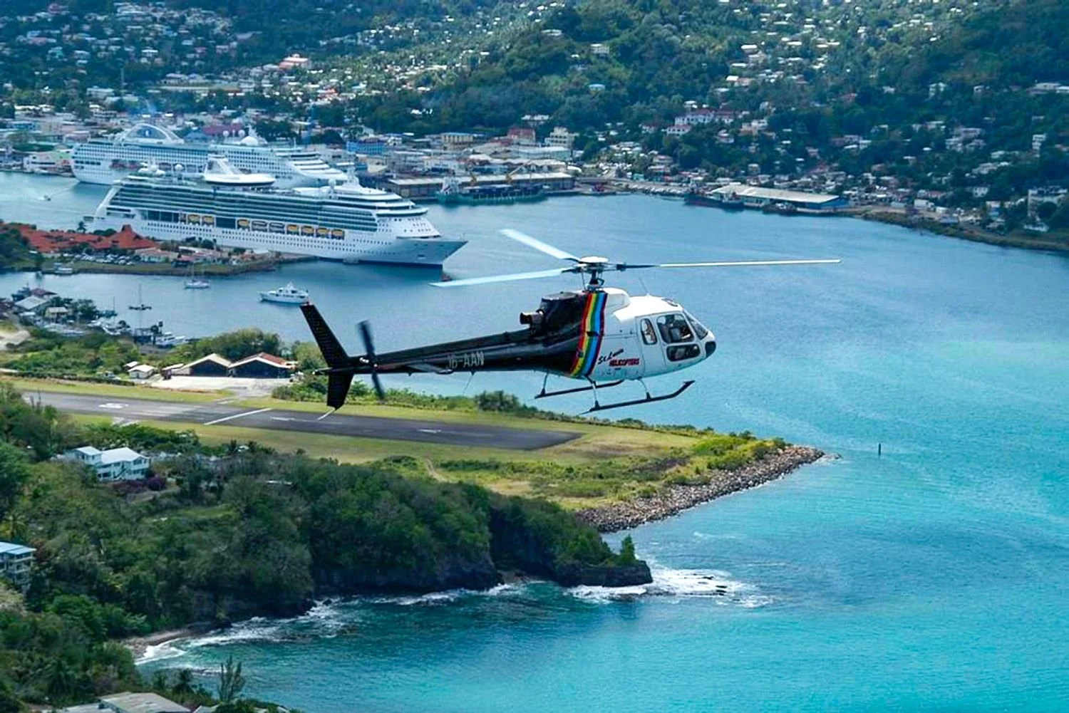 A helicopter flying over a coastal area with a harbor and large cruise ships docked nearby, surrounded by green hills and residential buildings.
