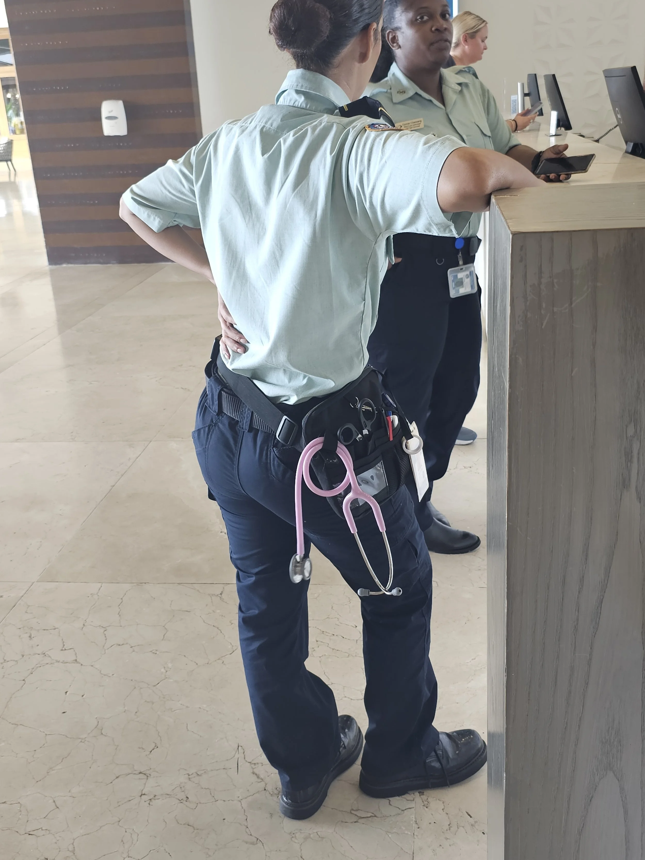 A female officer at a reception desk, wearing a uniform with a stethoscope hanging from her belt, engaged in conversation with another woman at the front counter.